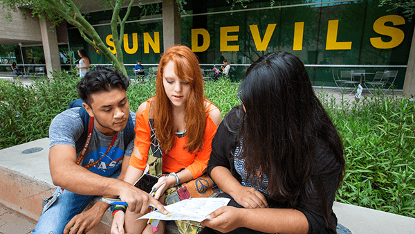 male student with gray NASA shirt and black hair, female student with orange shirt and red hair, and female student with black shirt and black hair looking at a piece of paper on campus