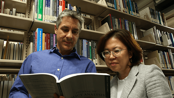 male professor with gray hair and blue collared shirt and female professor with brown hair, glasses, and gray blazer reading a book in the library
