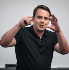 Dustin Pardini headshot - fair-skinned man, brown hair, hands raised near face, black collared shirt