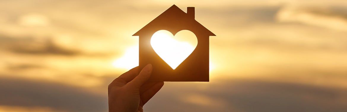hand holding a piece of paper in the shape of a house with a heart cutout against a sunset background