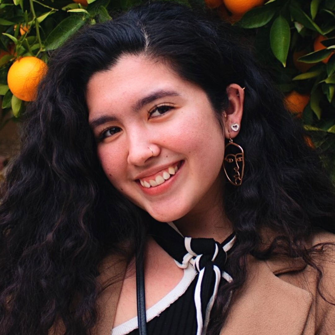 Young smiling woman with long, dark, curly hair and medium complexion wearing large earrings, black and white top, with tan coat over