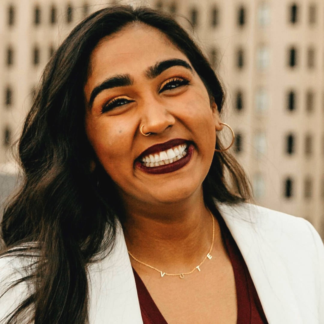Young woman with long dark hair and, with medium-dark complexion smiles a big smile. Wearing gold hoop earrings, a gold hoop nose ring, and dark lipstick. Dressed in a maroon top with white blazer standing on a balcony with skyscraper in the background