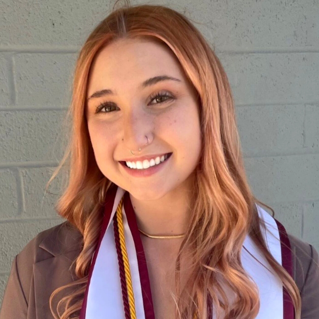 Young woman with light complexion and long, strawberry blonde hair smiles at camera wearing a stole and honor chords