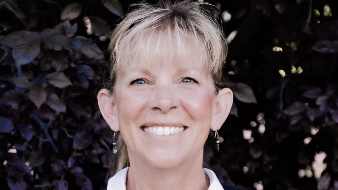 Kelly Bricker headshot - fair-skinned woman, smiling, blonde hair, white collared shirt, orange and black vest, earrings